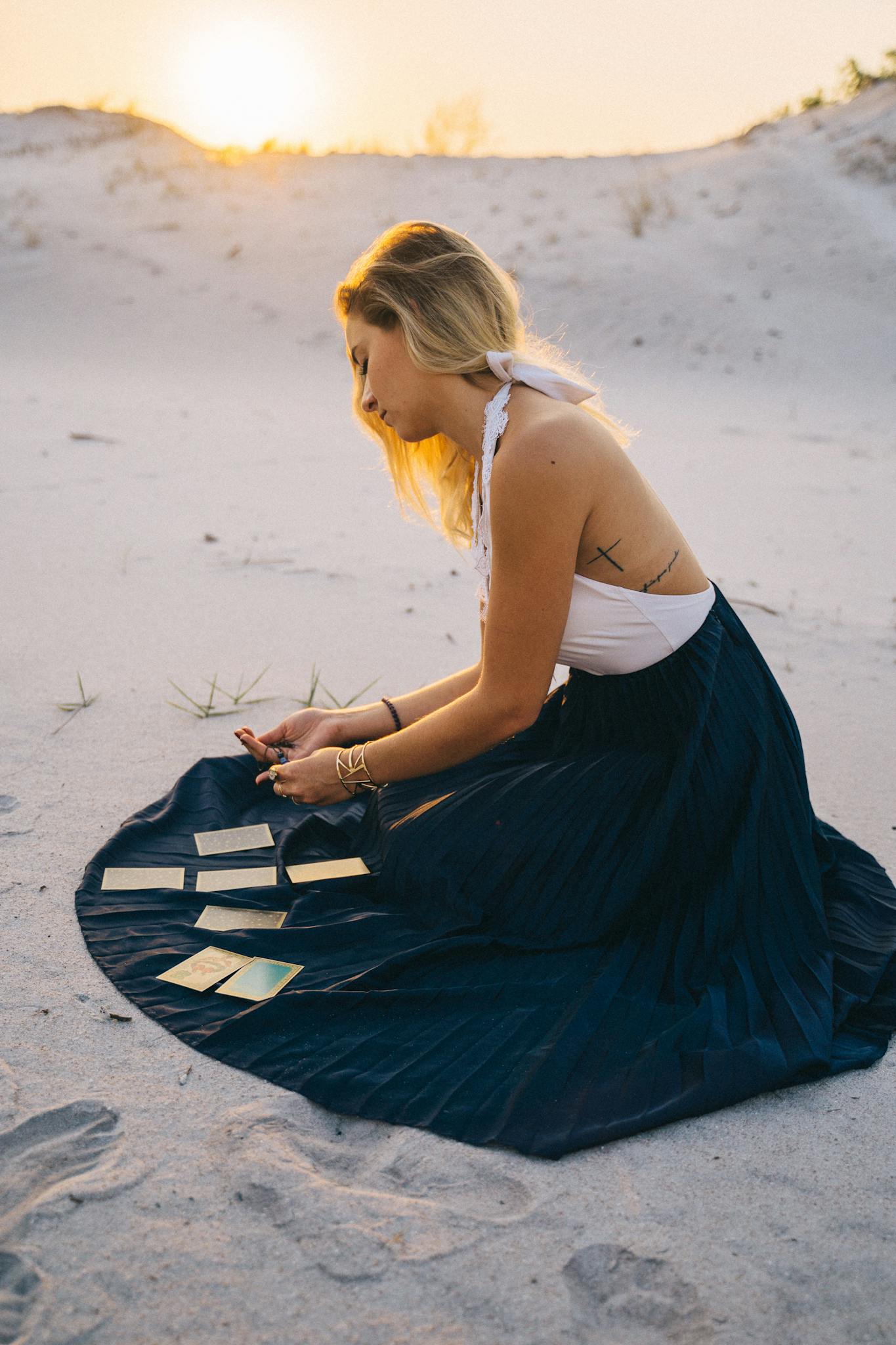 A woman sits on sand reading tarot cards during a desert sunset.